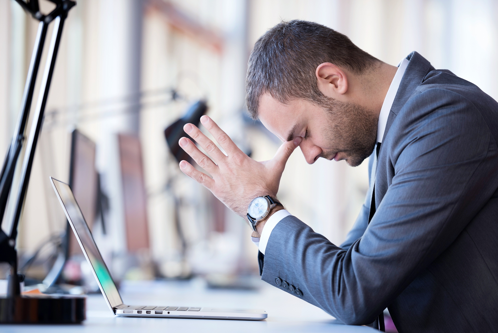 Man in business suit with closed eyes and head in hands in front of laptop worried about his dental website agency.